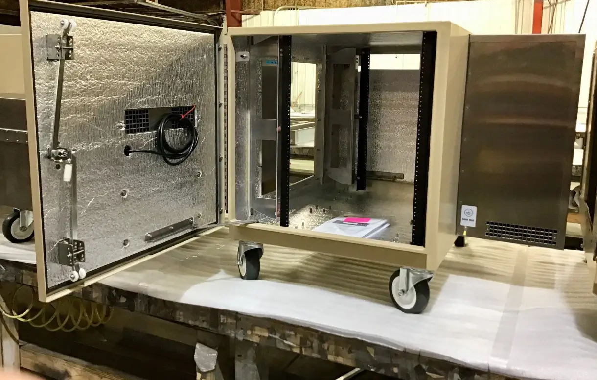 Large outdoor electrical enclosure with open doors showing insulated interior, rack mount rails, and caster wheels during fabrication